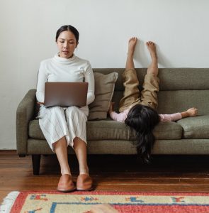 Woman working on a laptop on a sofa while a child plays beside her